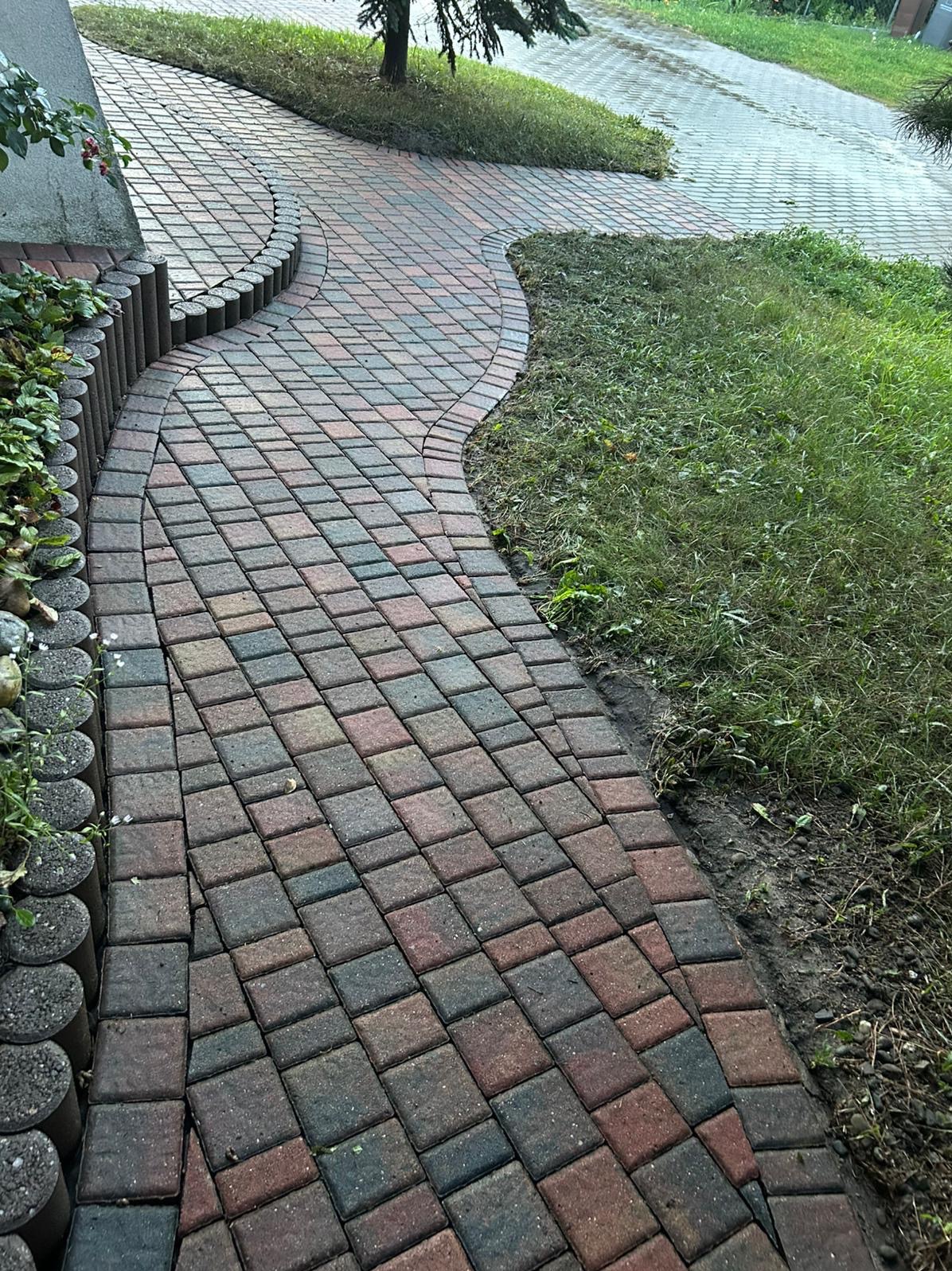 Kaskadowa ścieżka z kostki brukowej w odcieniach czerwieni i szarości, wijąca się przez trawnik, obrzeżona drewnianymi palisadami. Widok z góry, po deszczu.