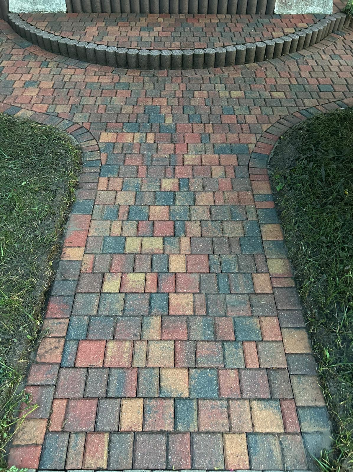 Kolorowa kostka brukowa ułożona w ścieżkę z obrzeżami, otoczona trawą. Widok z góry na wzór ułożenia kostki i jej barwy.