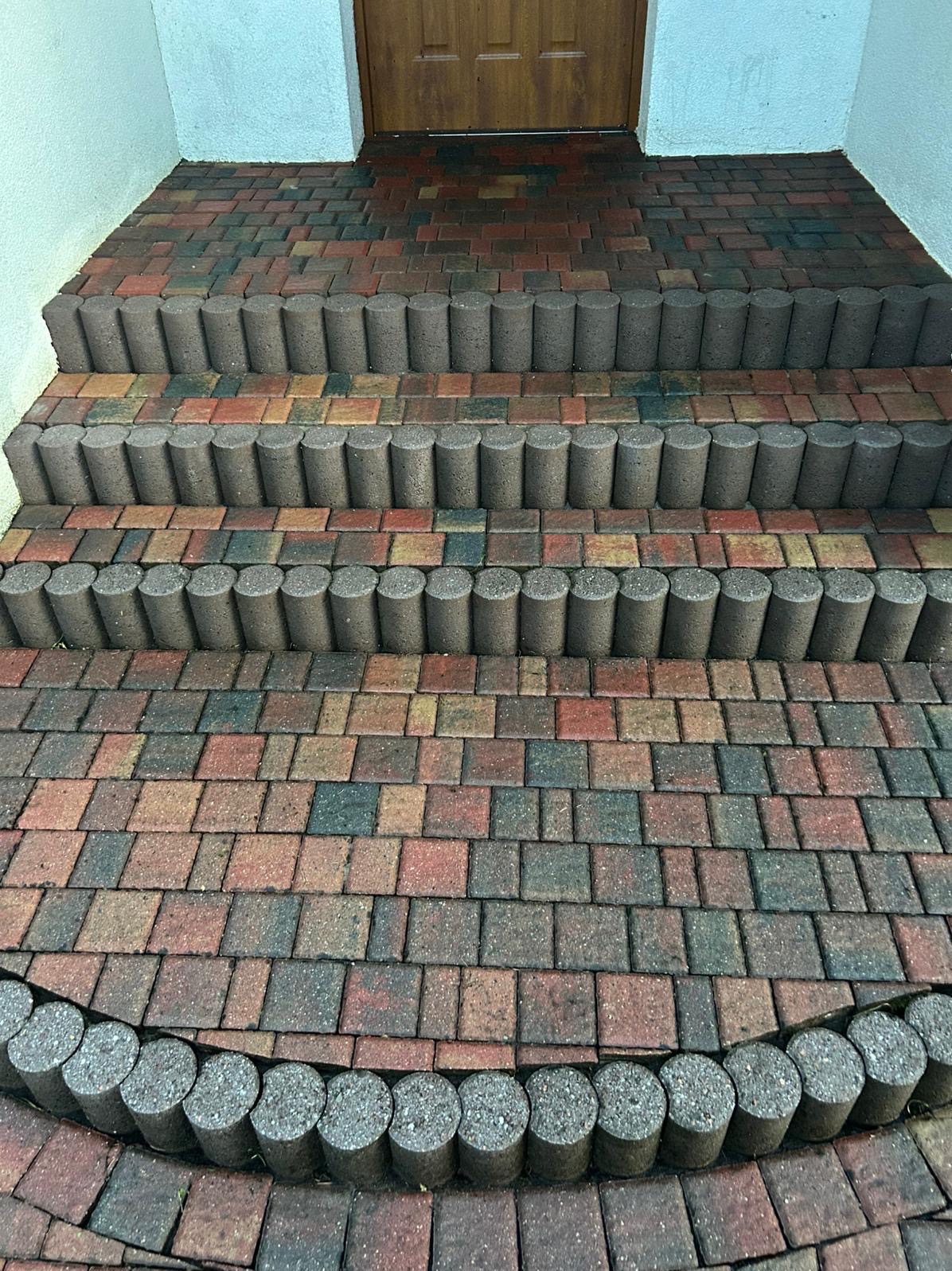 Schody wejściowe wyłożone kolorową kostką brukową i obrzeżami z betonowych palisad. Widoczne różnice w czystości kostki, sugerujące niedawne czyszczenie.