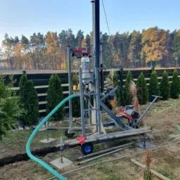 Wiertnica do studni głębinowych na działce, widoczny przewód doprowadzający wodę, w tle rząd tuj i las.