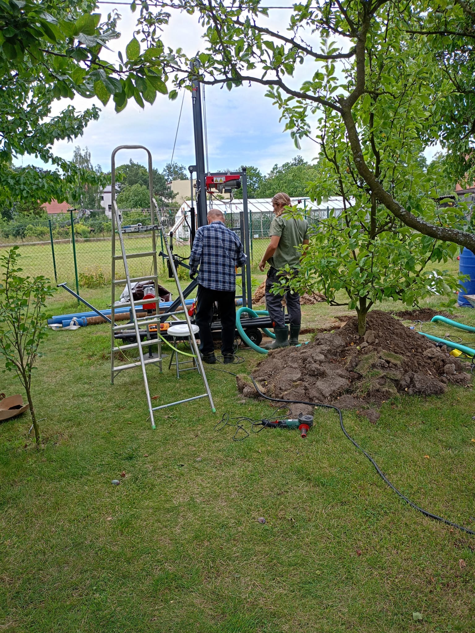 Dwóch mężczyzn przy maszynie wiertniczej na trawniku, widoczne narzędzia, drabina i wykopana ziemia.