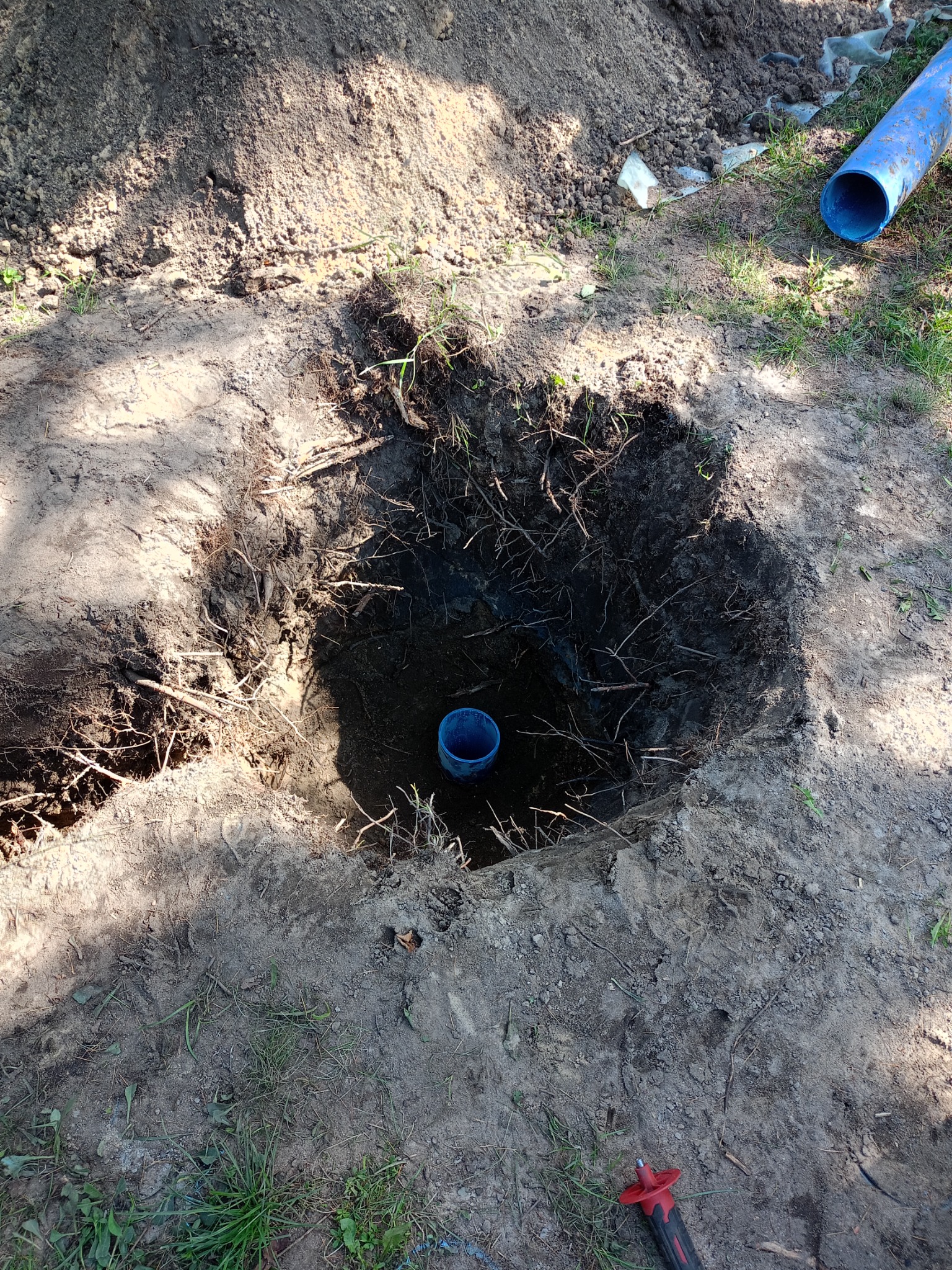Wykopany dół w ziemi z widocznymi korzeniami, na dnie niebieska rura; obok leży fragment niebieskiej rury i narzędzie z czerwoną rączką.