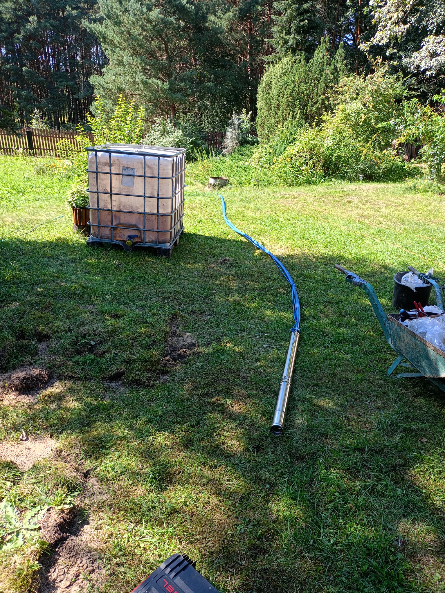 Pompa głębinowa z niebieskim przewodem leżąca na trawie obok zbiornika IBC i taczki z narzędziami na tle zieleni.