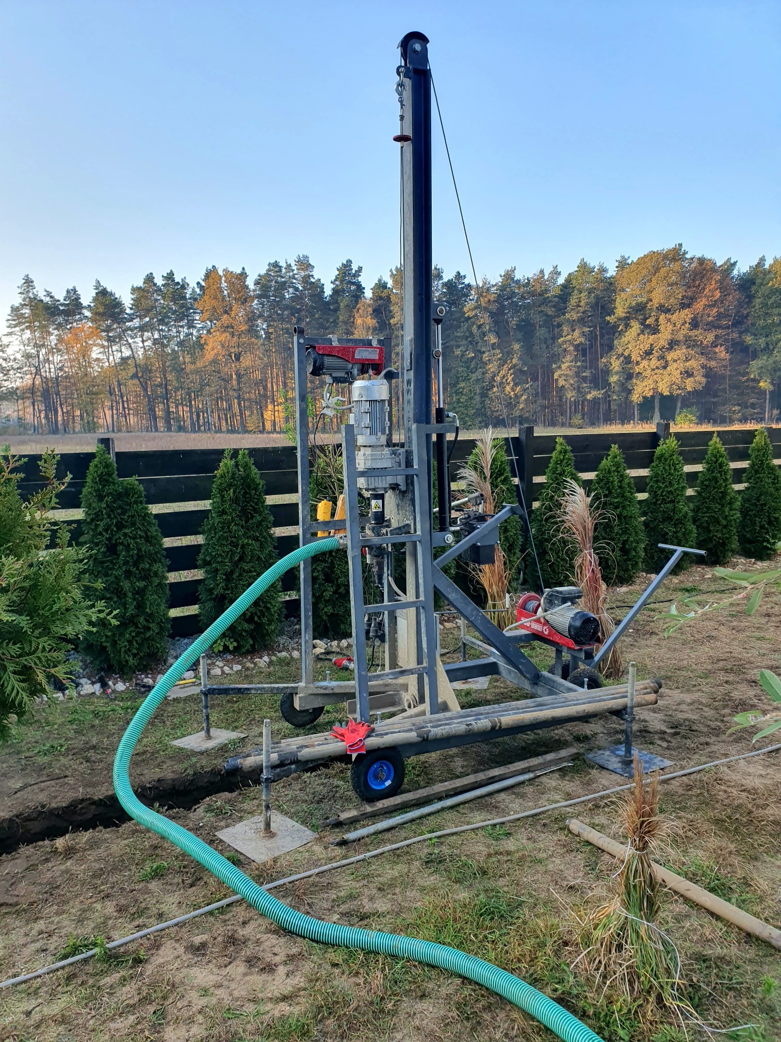 Wiertnica do studni głębinowych na działce, widoczny przewód doprowadzający wodę, w tle rząd tuj i las.