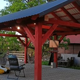 Dream-Projekt - Zadaszona, czerwona konstrukcja z drewnianych belek i metalowym dachem, oświetlona żarówkami, z drewnianą podłogą, hamakami, krzesłem i pufą w tle zieleni.