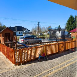 Dream-Projekt - Drewniane ogrodzenie placu zabaw z piaskiem, domkiem, zjeżdżalnią i trampoliną, widok z okna na podwórko z kostką brukową.