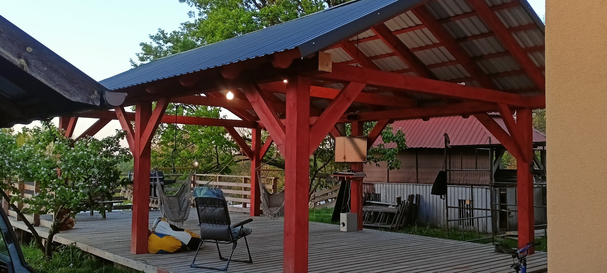 Zadaszona, czerwona konstrukcja z drewnianych belek i metalowym dachem, oświetlona żarówkami, z drewnianą podłogą, hamakami, krzesłem i pufą w tle zieleni.