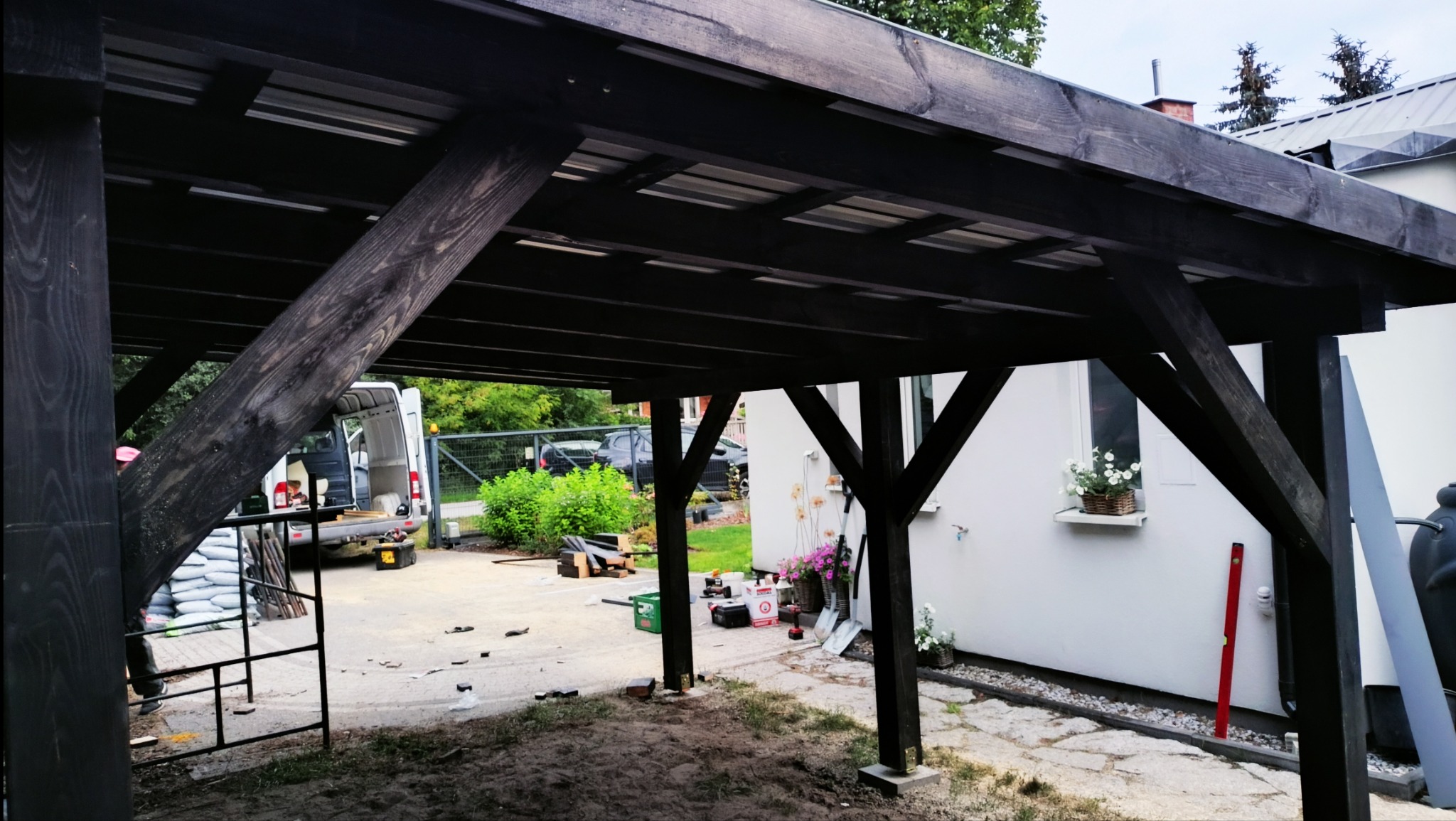 Budowa drewnianej pergoli w Łaziskach, widok od wewnątrz konstrukcji z widocznym samochodem dostawczym i materiałami budowlanymi, kwiaty w oknie domu.