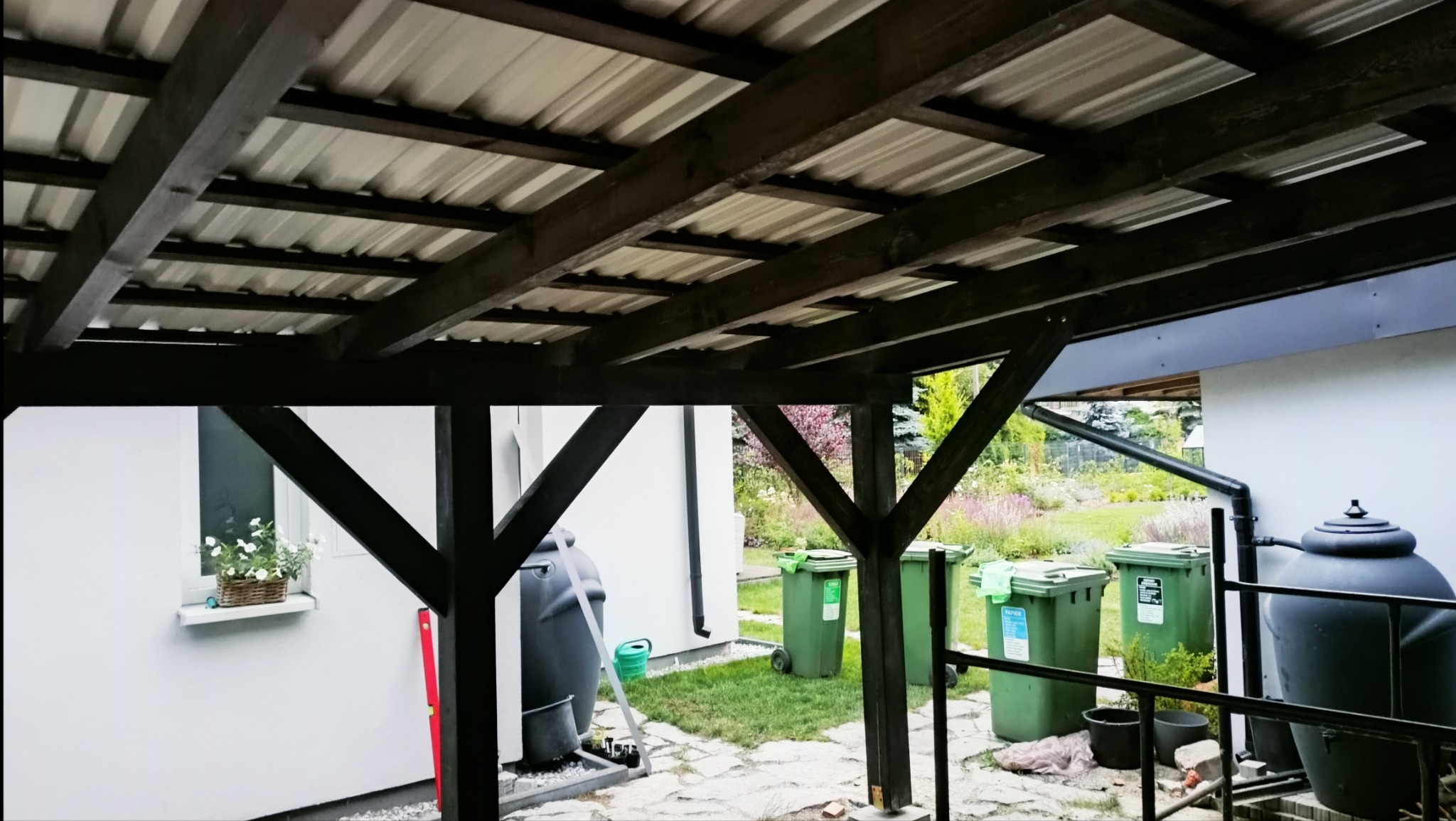 Zadaszenie tarasu z drewnianą konstrukcją, widok od dołu na metalowe poszycie dachu, w tle ogród z pojemnikami na śmieci i zbiornikiem na deszczówkę.