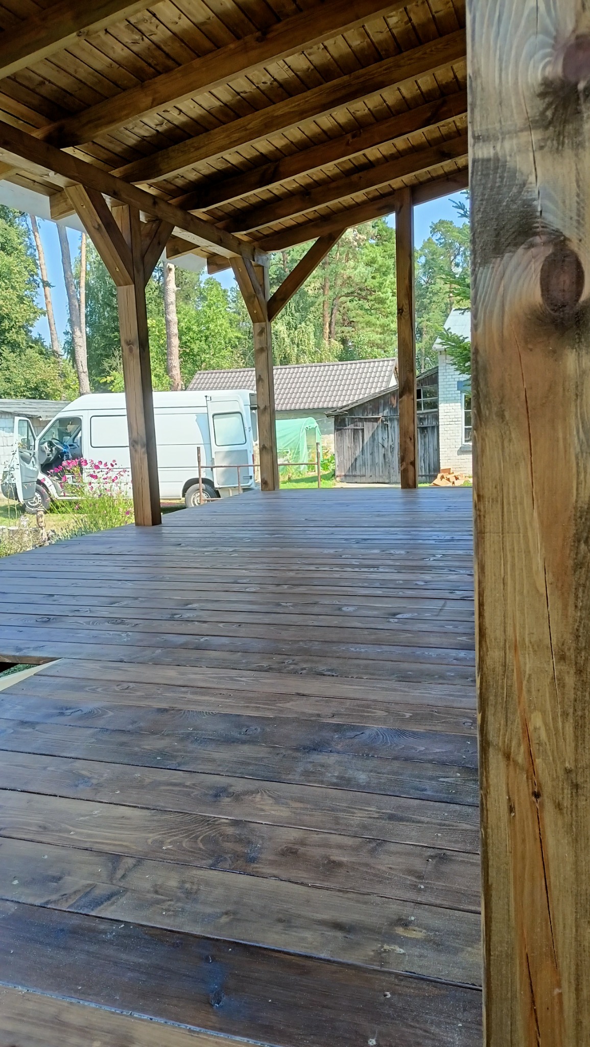 Zadaszony taras z drewnianą podłogą i belkami, widok na podwórze z zaparkowanym białym vanem w tle.