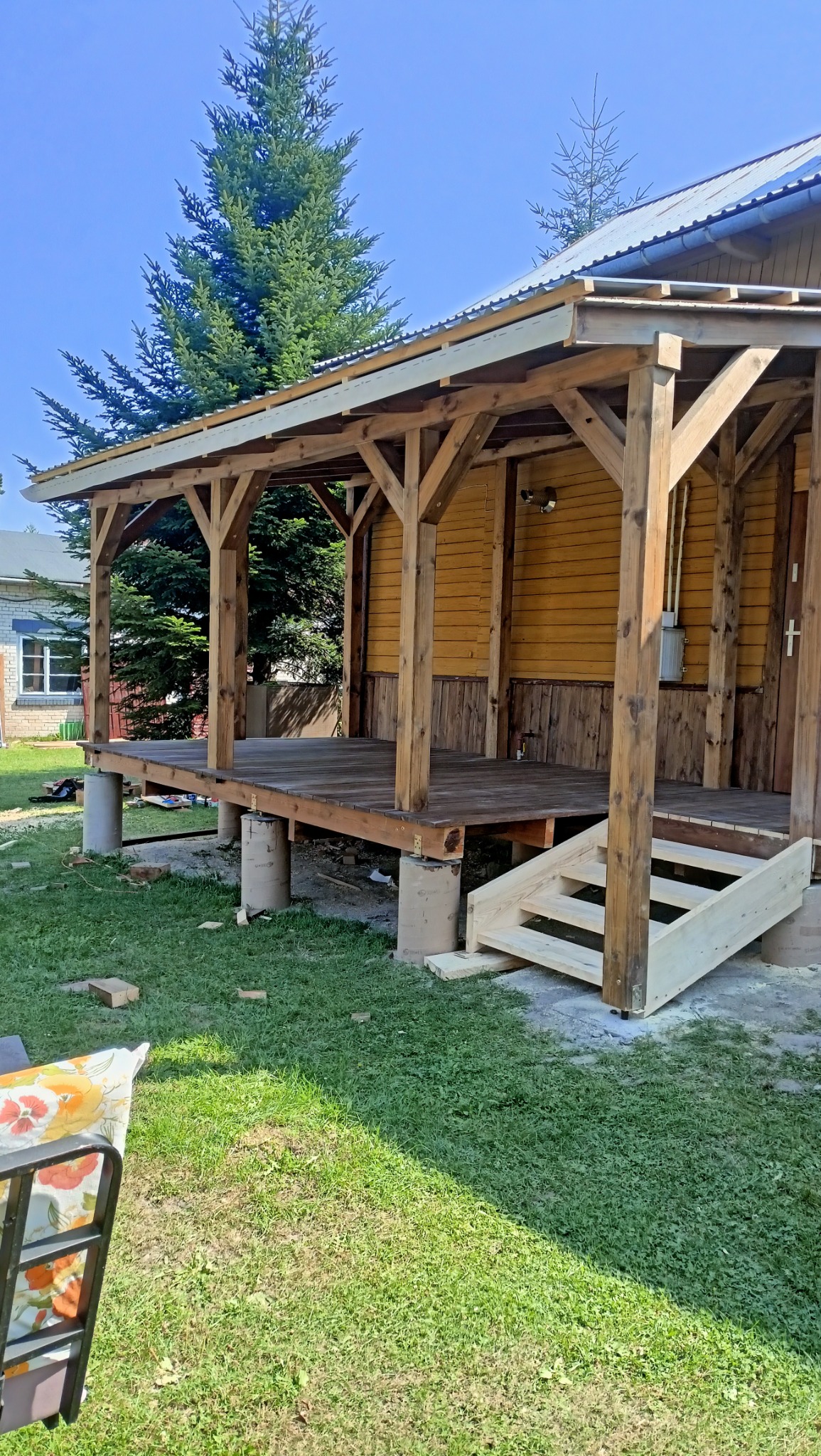 Drewniana konstrukcja z zadaszeniem na betonowych podstawach, częściowo wykończona, z surowymi schodami wejściowymi, na tle zieleni i budynku mieszkalnego.