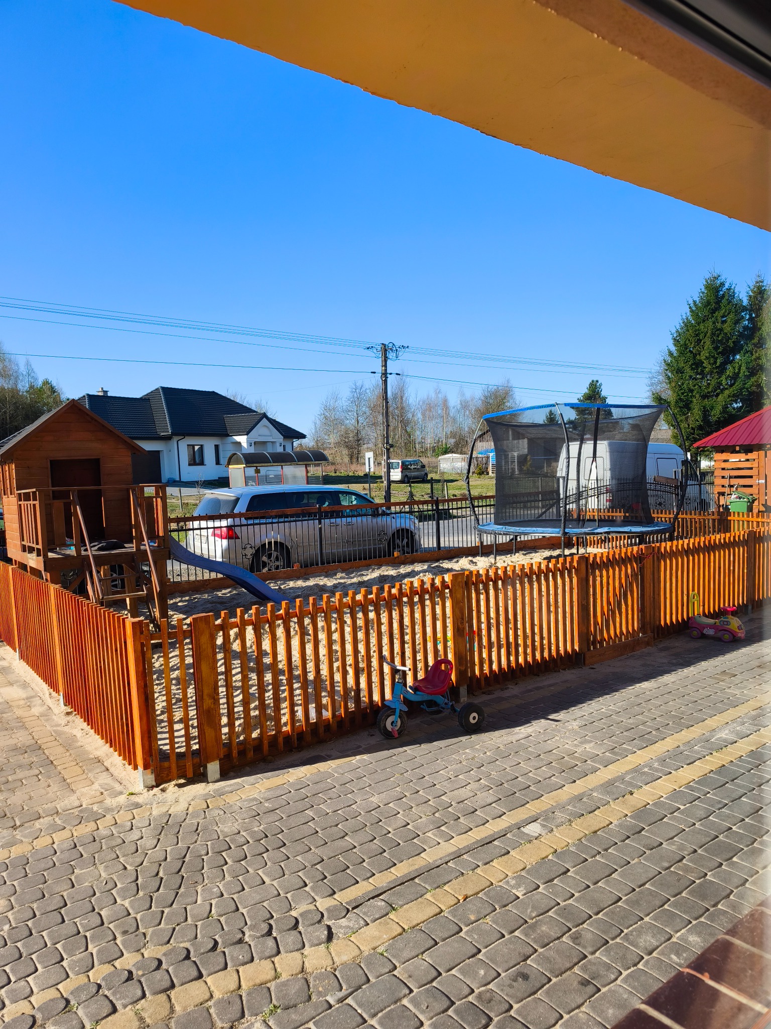 Drewniane ogrodzenie placu zabaw z piaskiem, domkiem, zjeżdżalnią i trampoliną, widok z okna na podwórko z kostką brukową.