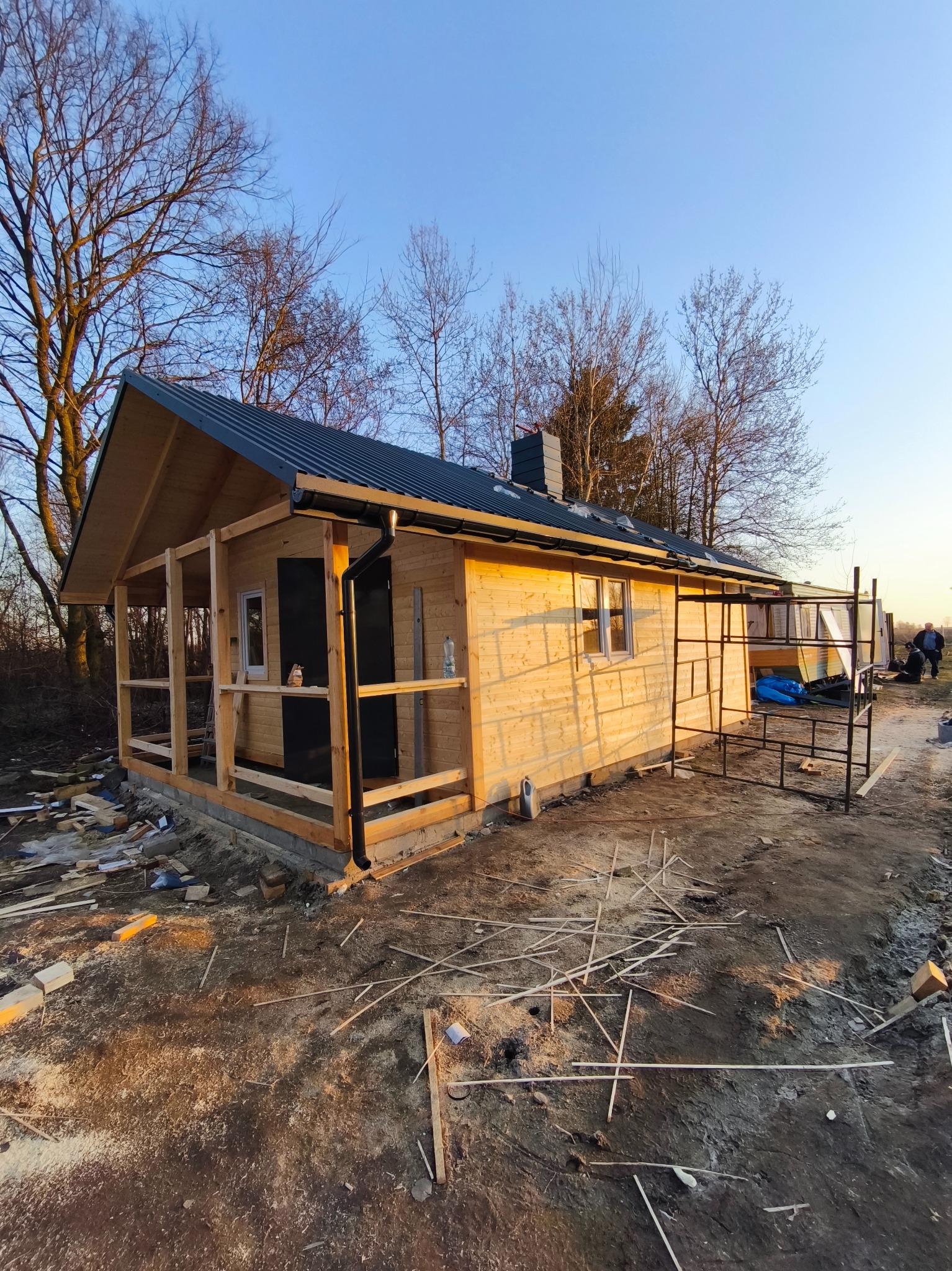 Budowa drewnianej konstrukcji z czarnym dachem i kominem, widoczne rusztowania i elementy konstrukcyjne, teren budowy w trakcie prac.