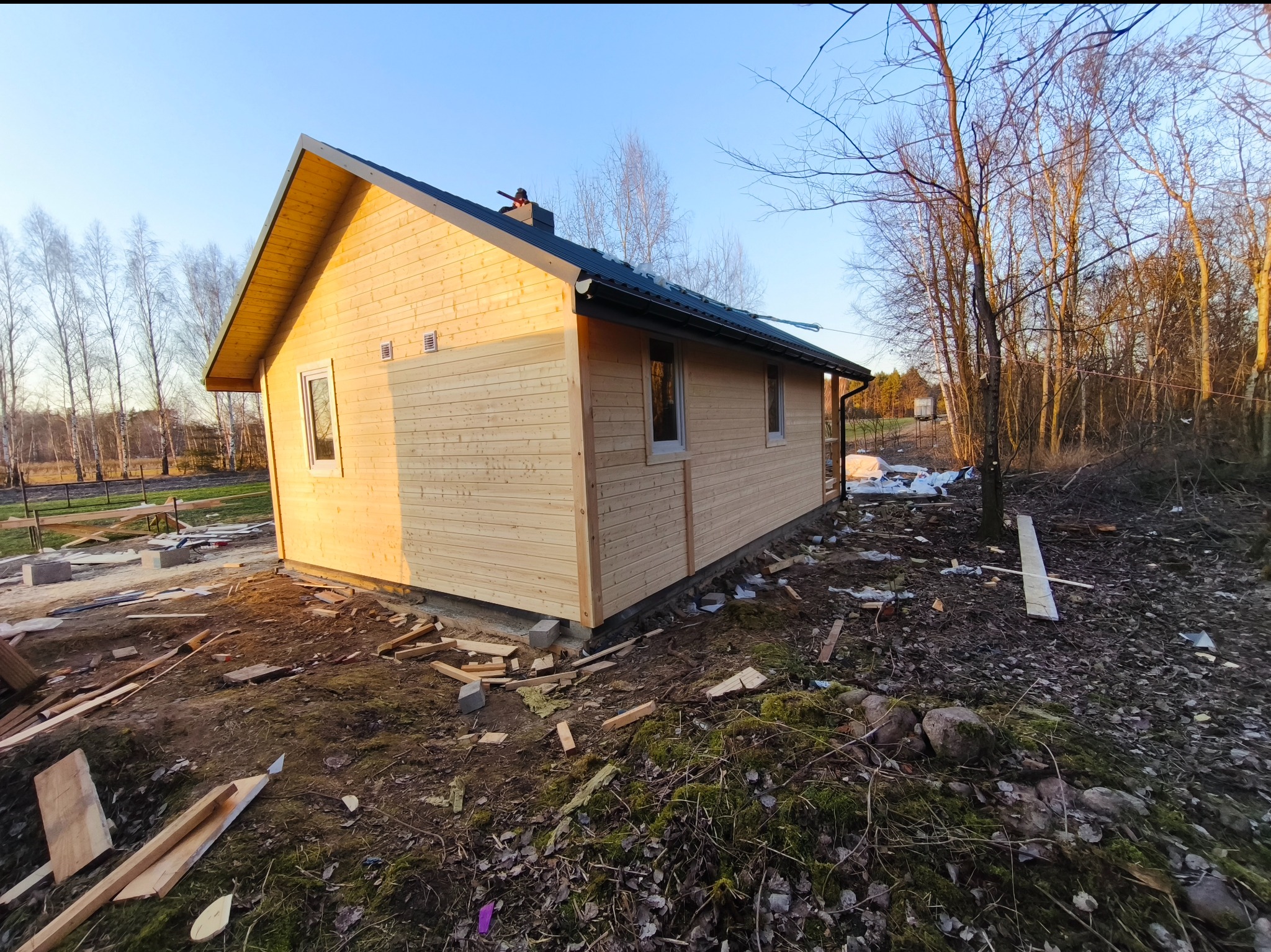 Surowa, drewniana konstrukcja domu w trakcie budowy, z widocznymi elementami izolacji i odpadami budowlanymi na ziemi, kontrastująca z otaczającym lasem i błękitnym niebem.