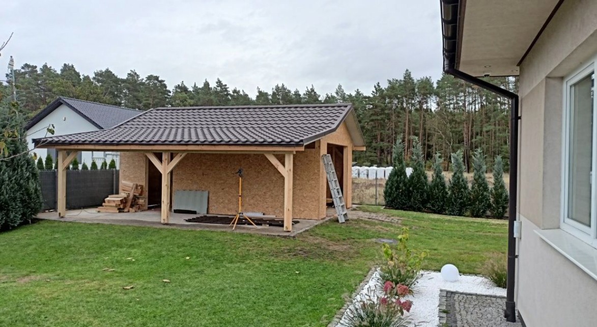 Budowa zadaszonej konstrukcji z drewnianymi słupami i brązowym dachem na posesji, widoczne materiały budowlane, drabina i lampa robocza.