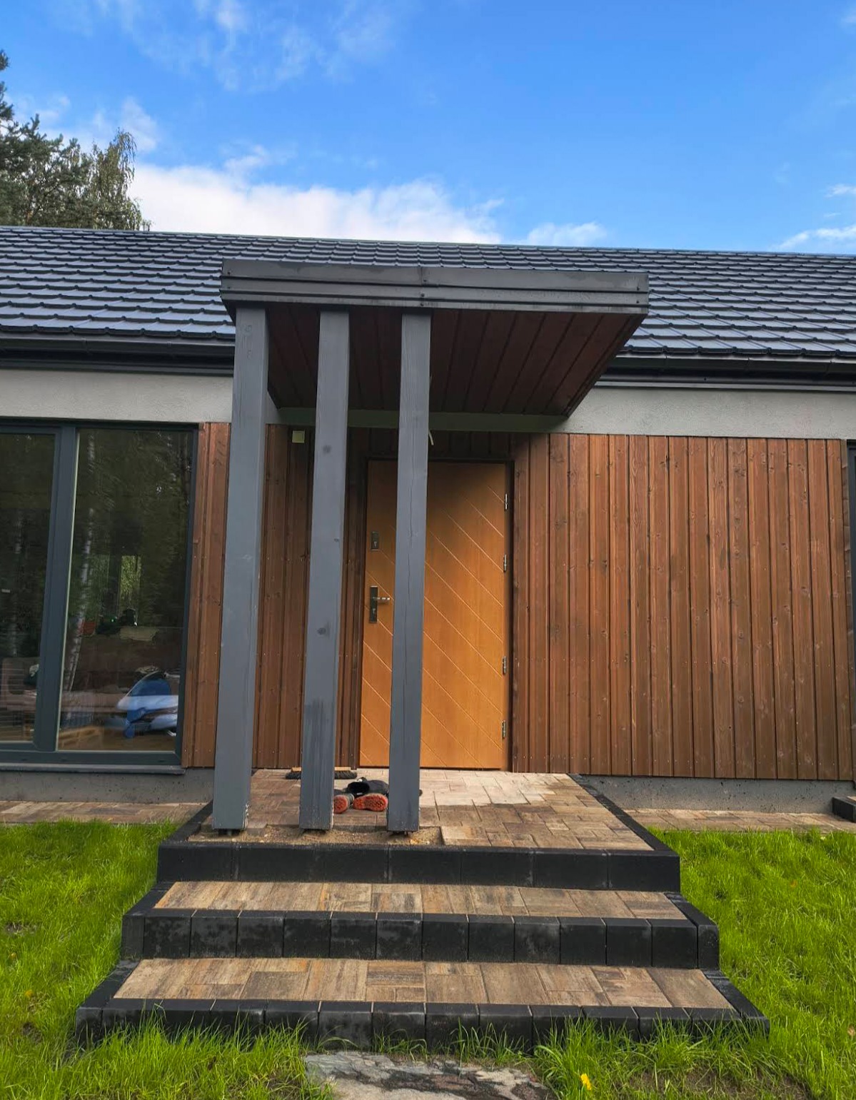Nowoczesne wejście do domu z drewnianą elewacją i zadaszeniem wspartym na szarych słupach. Schody z kostki brukowej prowadzą do drzwi. Widoczne narzędzia.