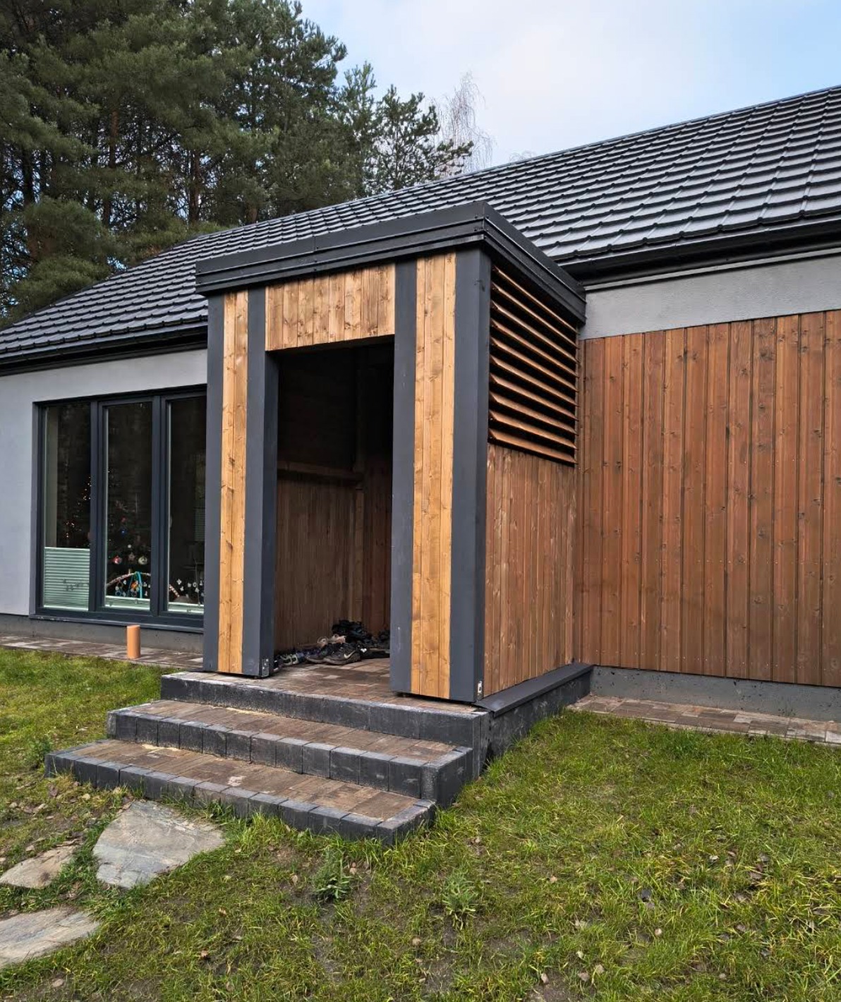 Drewniana weranda z szarymi elementami konstrukcyjnymi, prowadząca do domu z szarą dachówką. Widoczne schody z kostki brukowej i trawnik.