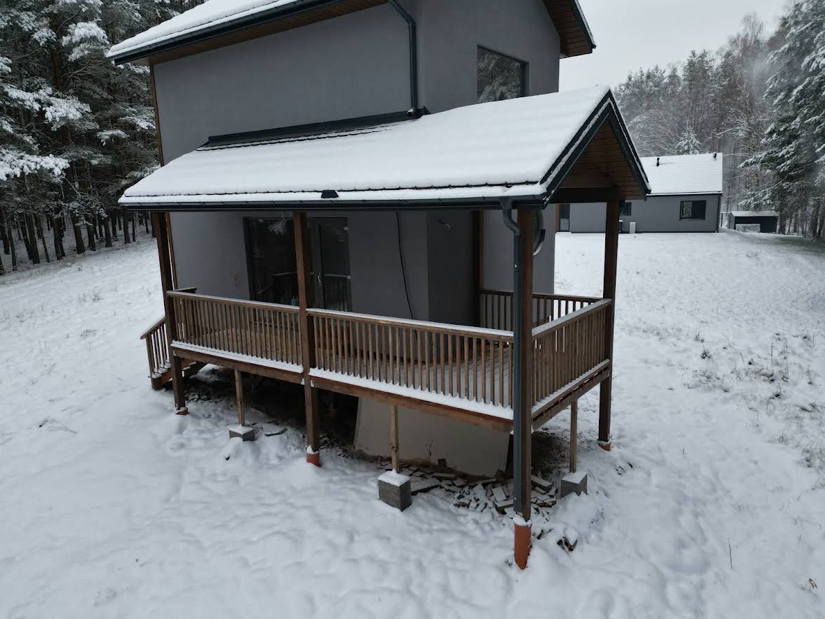 Zimowy krajobraz z drewnianym tarasem przy szarym domu. Śnieg pokrywa konstrukcję i otoczenie, tworząc spokojną, chłodną scenerię. Widok z góry.