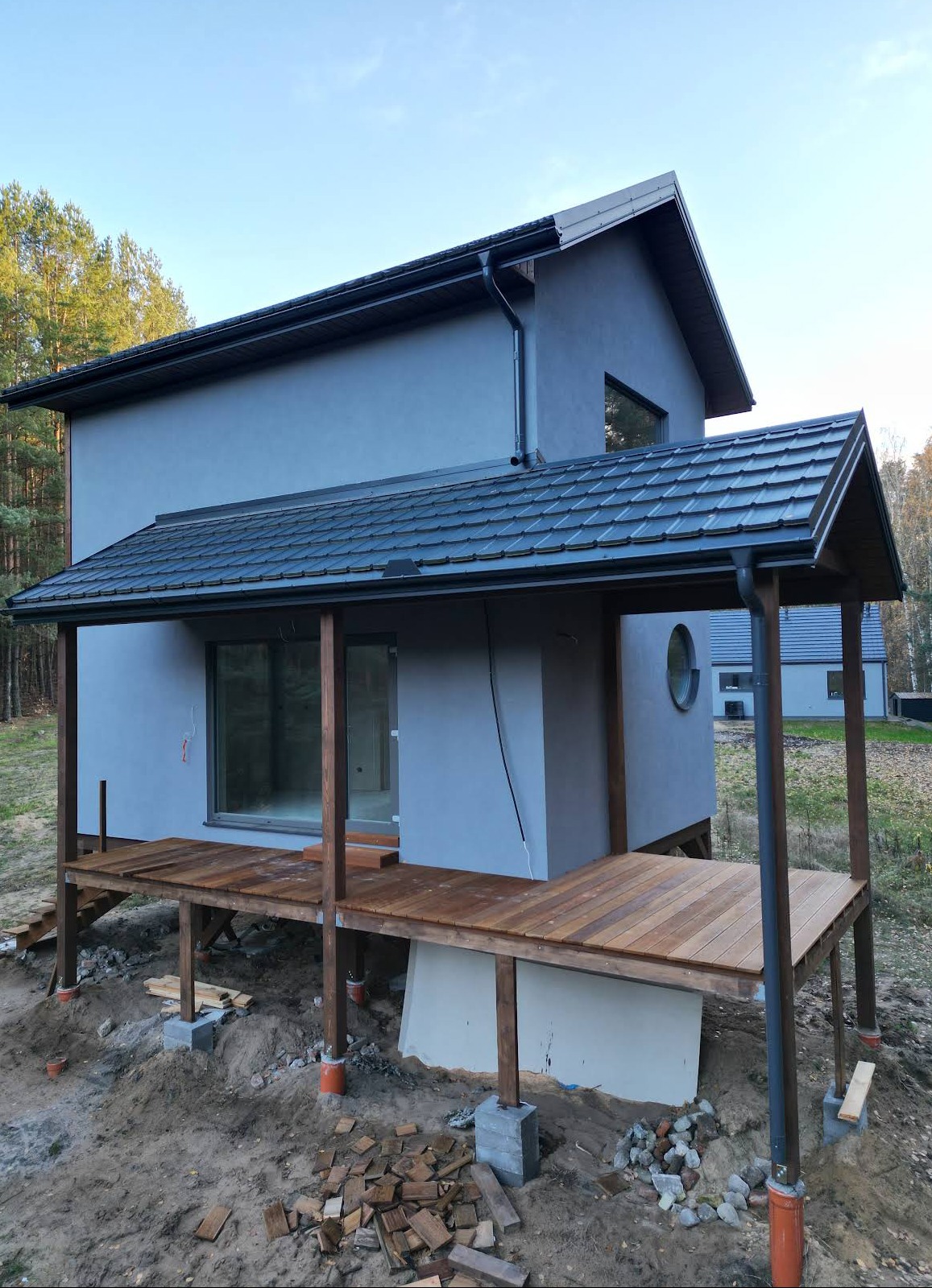 Nowoczesny dom z drewnianym tarasem w trakcie budowy, widoczne elementy konstrukcyjne i materiały budowlane. Surowy stan, detale wykończenia w trakcie realizacji.