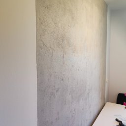 Usługi ogólnobudowlane Jakub Bochnia - Ściana wewnętrzna z dekoracyjną, szarą strukturą obok gładkiej, białej ściany. Widoczny fragment białego stołu z kluczami i telefonem.