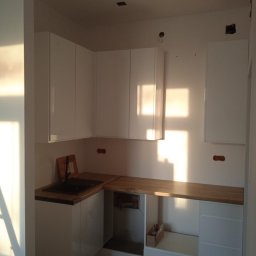 Usługi ogólnobudowlane Jakub Bochnia - Jasna kuchnia z białymi szafkami i drewnianym blatem, widoczne niedokończone prace instalacyjne na ścianie, promienie słoneczne wpadające przez okno.