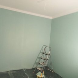 Usługi ogólnobudowlane Jakub Bochnia - Pomieszczenie po świeżym pomalowaniu ścian na pastelowy zielony kolor, widoczna drabina aluminiowa i wiaderko z farbą w rogu, podłoga zabezpieczona folią.
