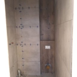 Usługi ogólnobudowlane Jakub Bochnia - Łazienka w trakcie wykańczania: ułożone płytki ścienne w odcieniach szarości i brązu, z widocznymi krzyżykami dystansowymi i poziomnicą na podłodze. Częściowo zamontowany stelaż podtynkowy WC.