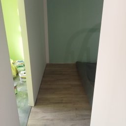 Usługi ogólnobudowlane Jakub Bochnia - Fragment wnętrza po remoncie: panele podłogowe ułożone, ściany pomalowane na zielono i biało, widoczne puszki z farbą w sąsiednim pomieszczeniu.