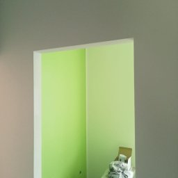 Usługi ogólnobudowlane Jakub Bochnia - Fragment odnowionego wnętrza z pomalowanymi na jasnozielono ścianami, widoczny przez otwór w ścianie pomalowanej na szaro, na pierwszym planie widoczne narzędzia malarskie i materiały budowlane.