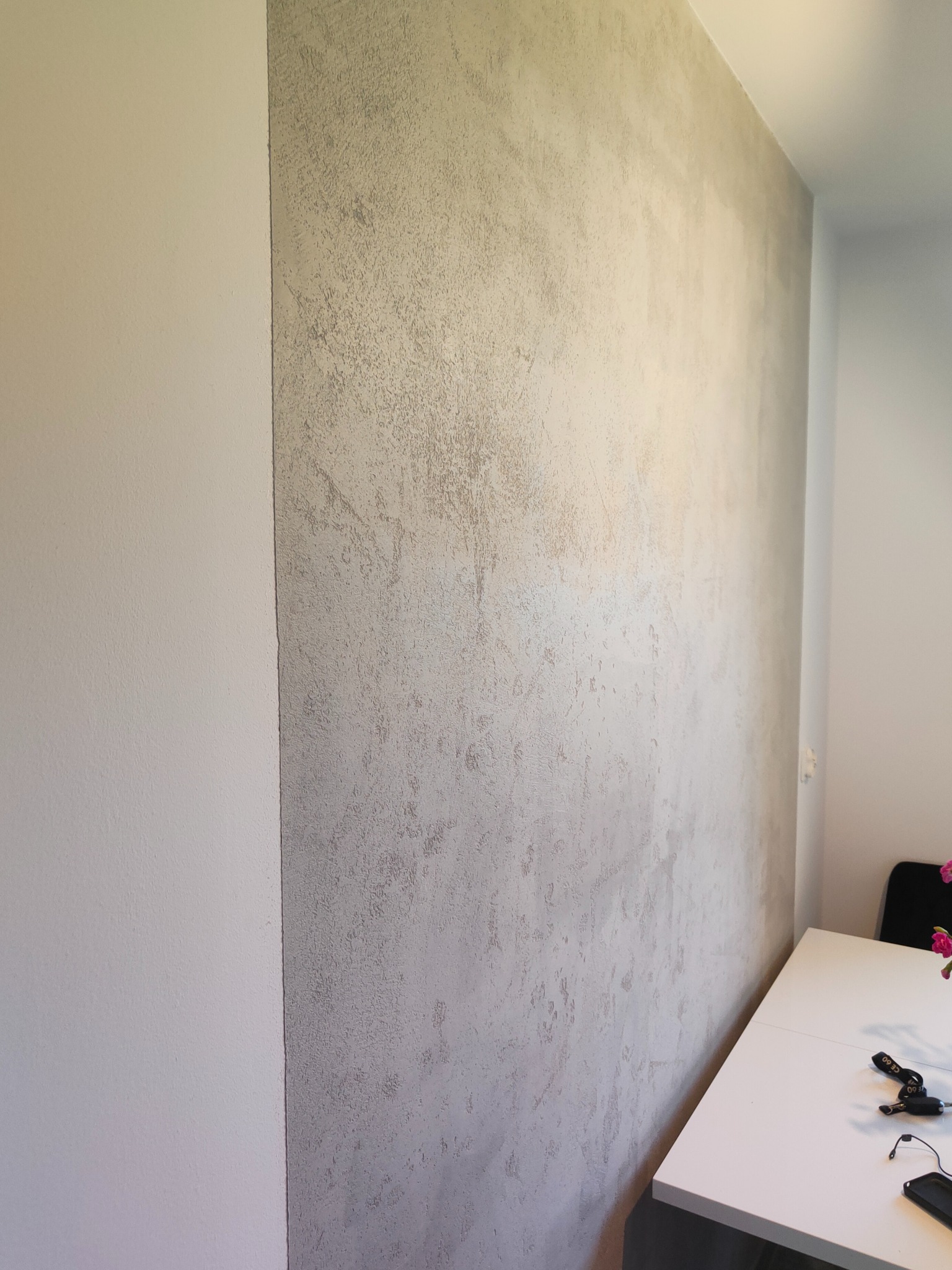 Ściana wewnętrzna z dekoracyjną, szarą strukturą obok gładkiej, białej ściany. Widoczny fragment białego stołu z kluczami i telefonem.