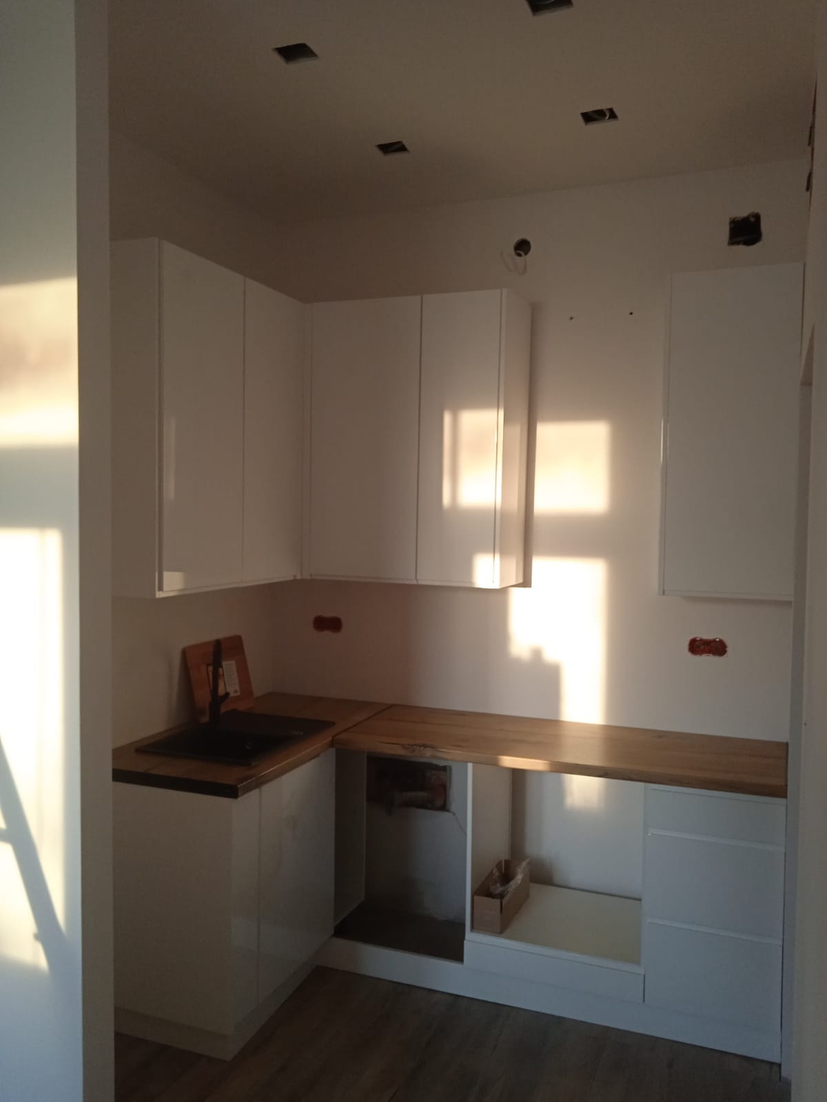 Jasna kuchnia z białymi szafkami i drewnianym blatem, widoczne niedokończone prace instalacyjne na ścianie, promienie słoneczne wpadające przez okno.