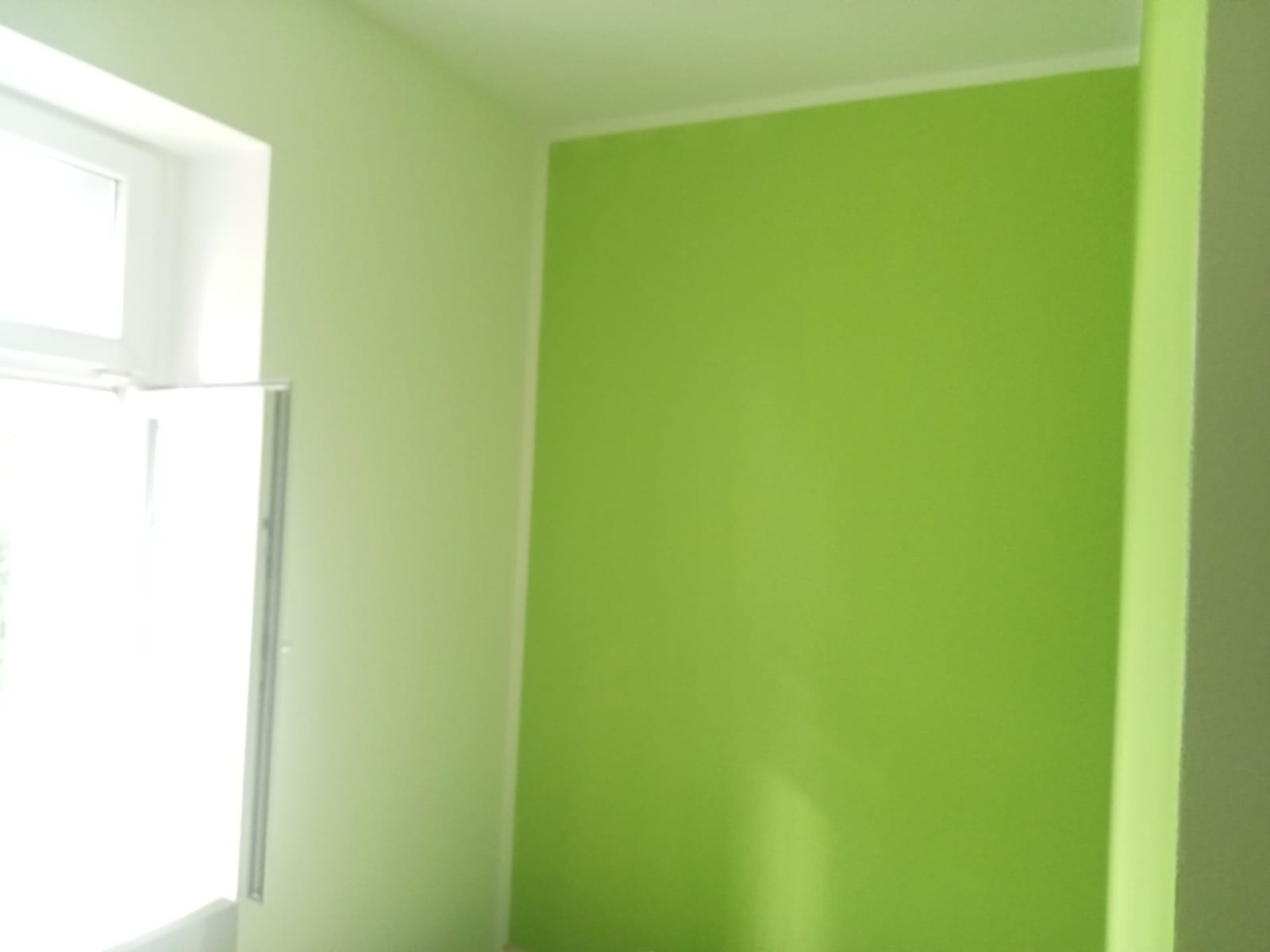 Pomalowany na zielono pokój z oknem po lewej stronie, widoczny fragment sufitu i listwa przysufitowa.