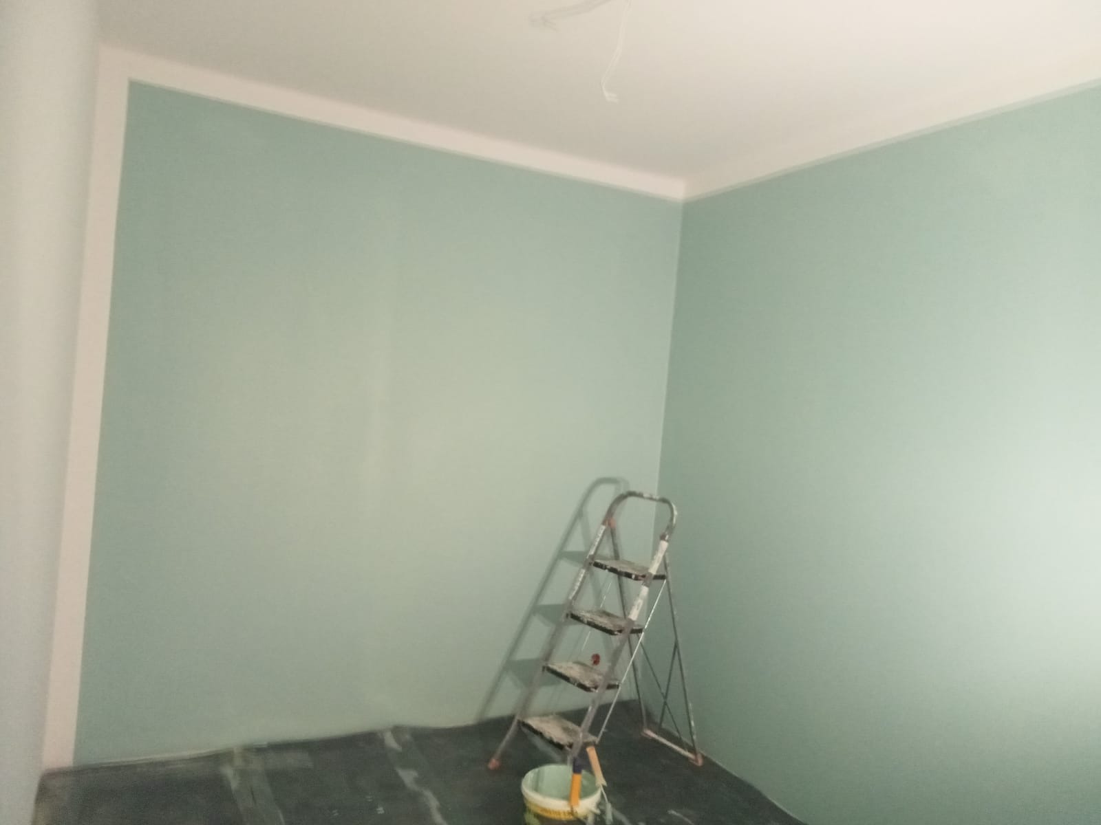 Pomieszczenie po świeżym pomalowaniu ścian na pastelowy zielony kolor, widoczna drabina aluminiowa i wiaderko z farbą w rogu, podłoga zabezpieczona folią.