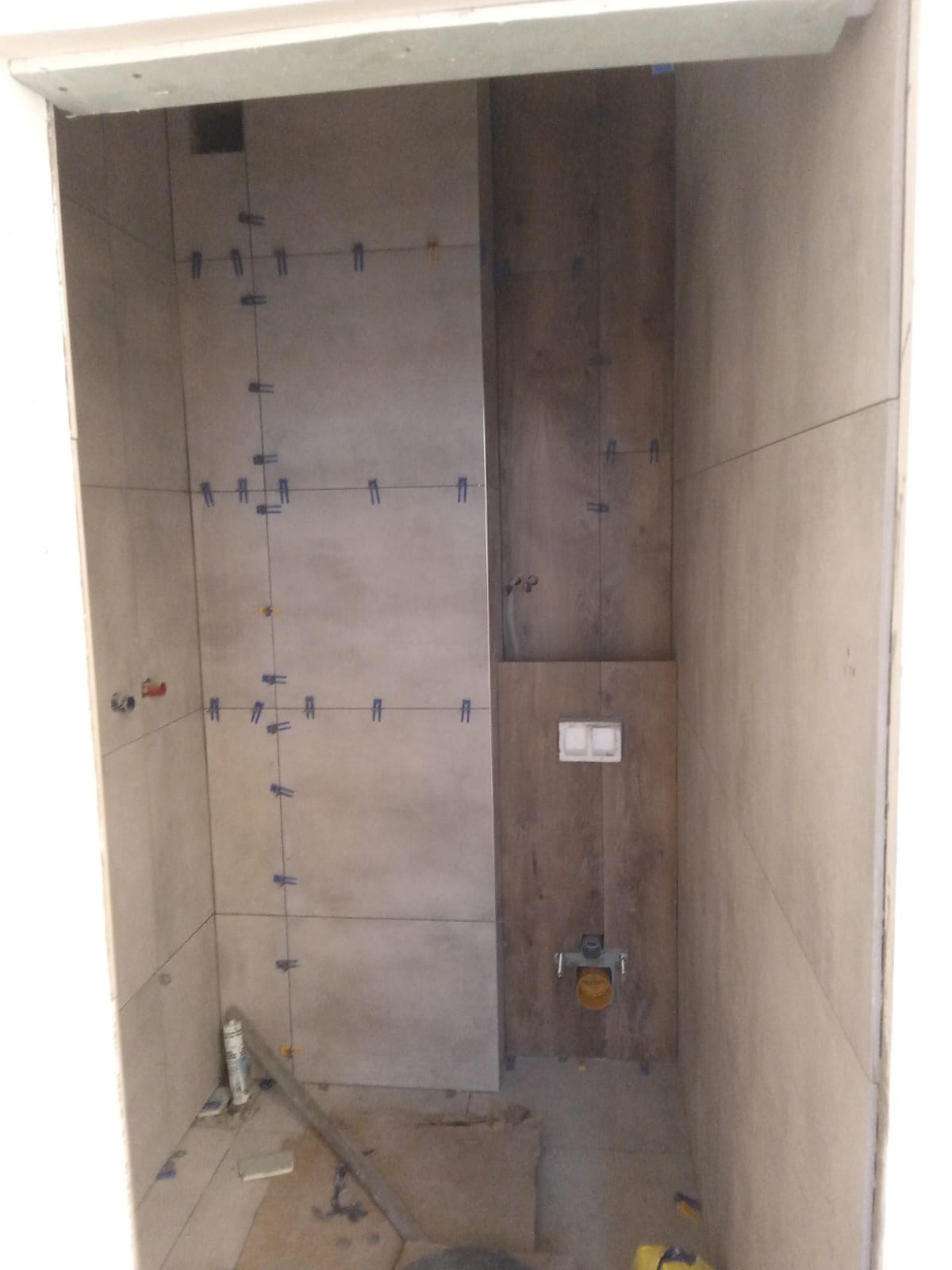 Łazienka w trakcie wykańczania: ułożone płytki ścienne w odcieniach szarości i brązu, z widocznymi krzyżykami dystansowymi i poziomnicą na podłodze. Częściowo zamontowany stelaż podtynkowy WC.