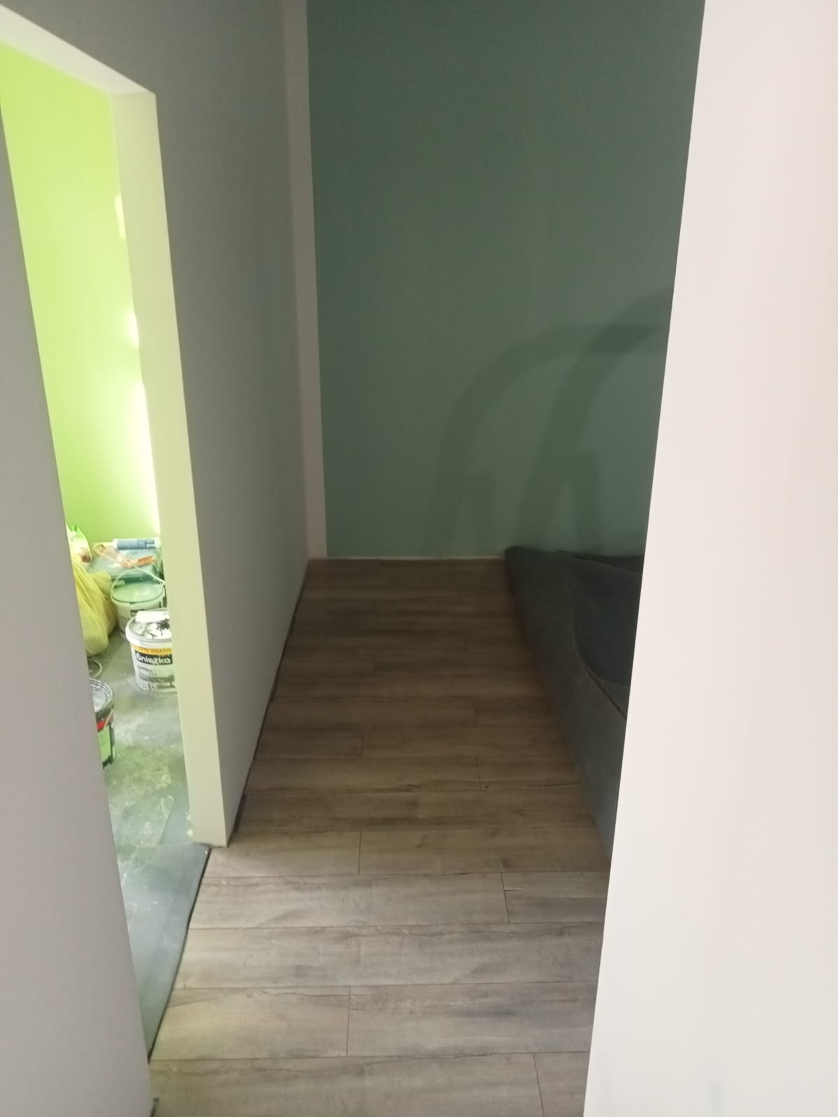 Fragment wnętrza po remoncie: panele podłogowe ułożone, ściany pomalowane na zielono i biało, widoczne puszki z farbą w sąsiednim pomieszczeniu.