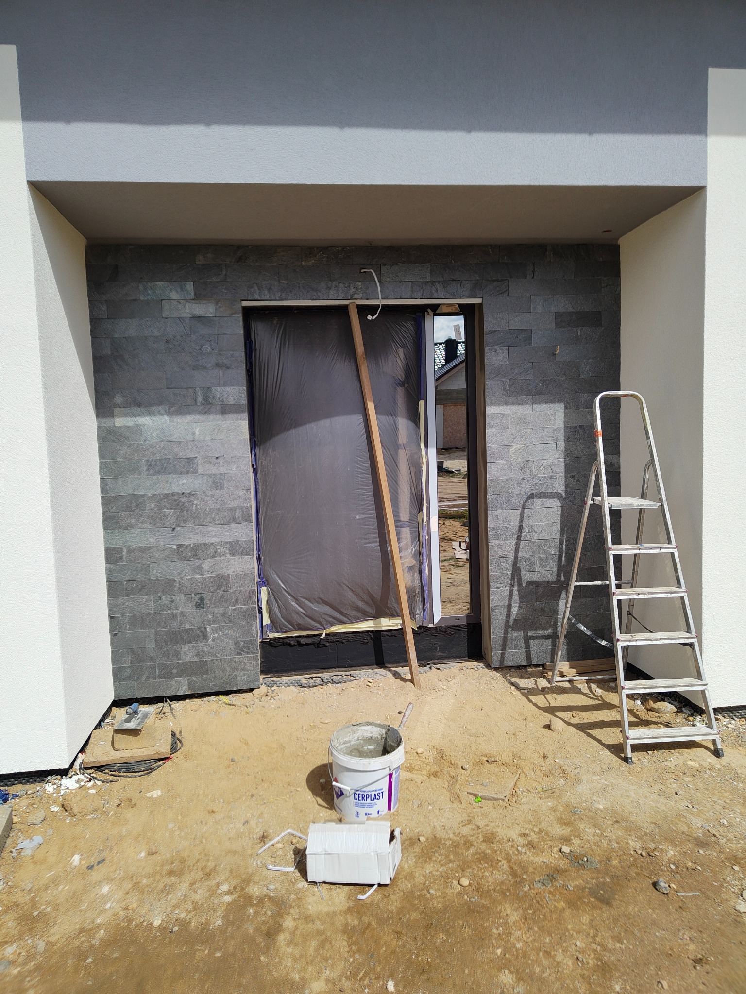 Montaż kamiennej okładziny elewacyjnej przy wejściu do budynku, zabezpieczone folią drzwi, drabina i wiadro z zaprawą na placu budowy.
