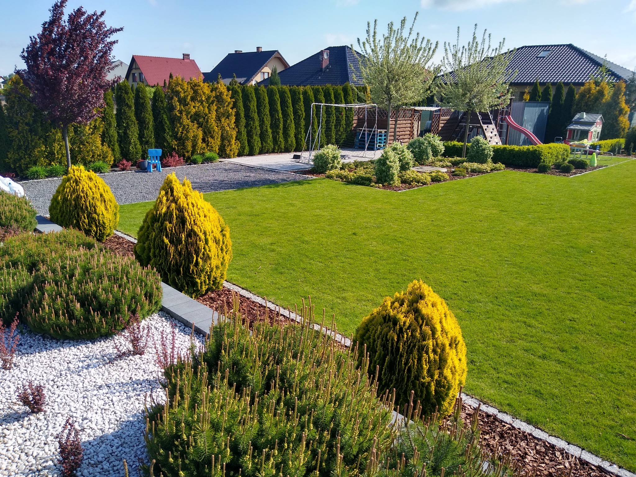 Starannie utrzymany ogród z równo przystrzyżonym trawnikiem, ozdobnymi krzewami, drzewami i wyżwirowanymi ścieżkami, w tle domy jednorodzinne i plac zabaw