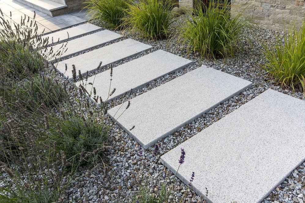 Ścieżka z prostokątnych, jasnoszarych płyt betonowych ułożonych w trawie z dekoracyjnym żwirem, obok rosną kępy lawendy i trawy ozdobnej.