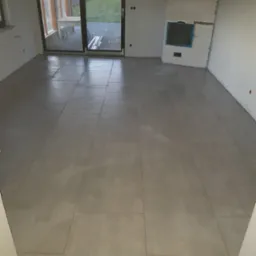 Ułożone szare płytki podłogowe w surowym wnętrzu z widokiem na zewnątrz przez duże przesuwne drzwi balkonowe.