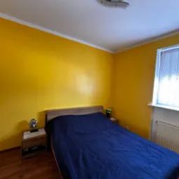 Sypialnia z intensywnie żółtymi ścianami, drewnianą podłogą i niebieską narzutą na łóżku; na szafce nocnej dekoracja w kształcie konia.