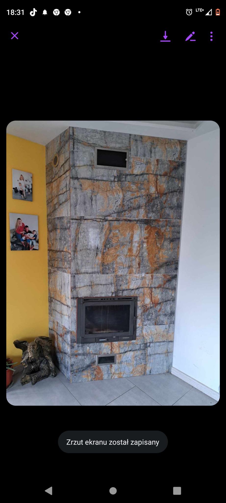 Obudowa kominka wykończona płytkami imitującymi marmur w odcieniach szarości i brązu, z wkładem kominkowym, wentylacją oraz dekoracyjną rzeźbą drewnianą na podłodze z szarych płytek, na ścianie...