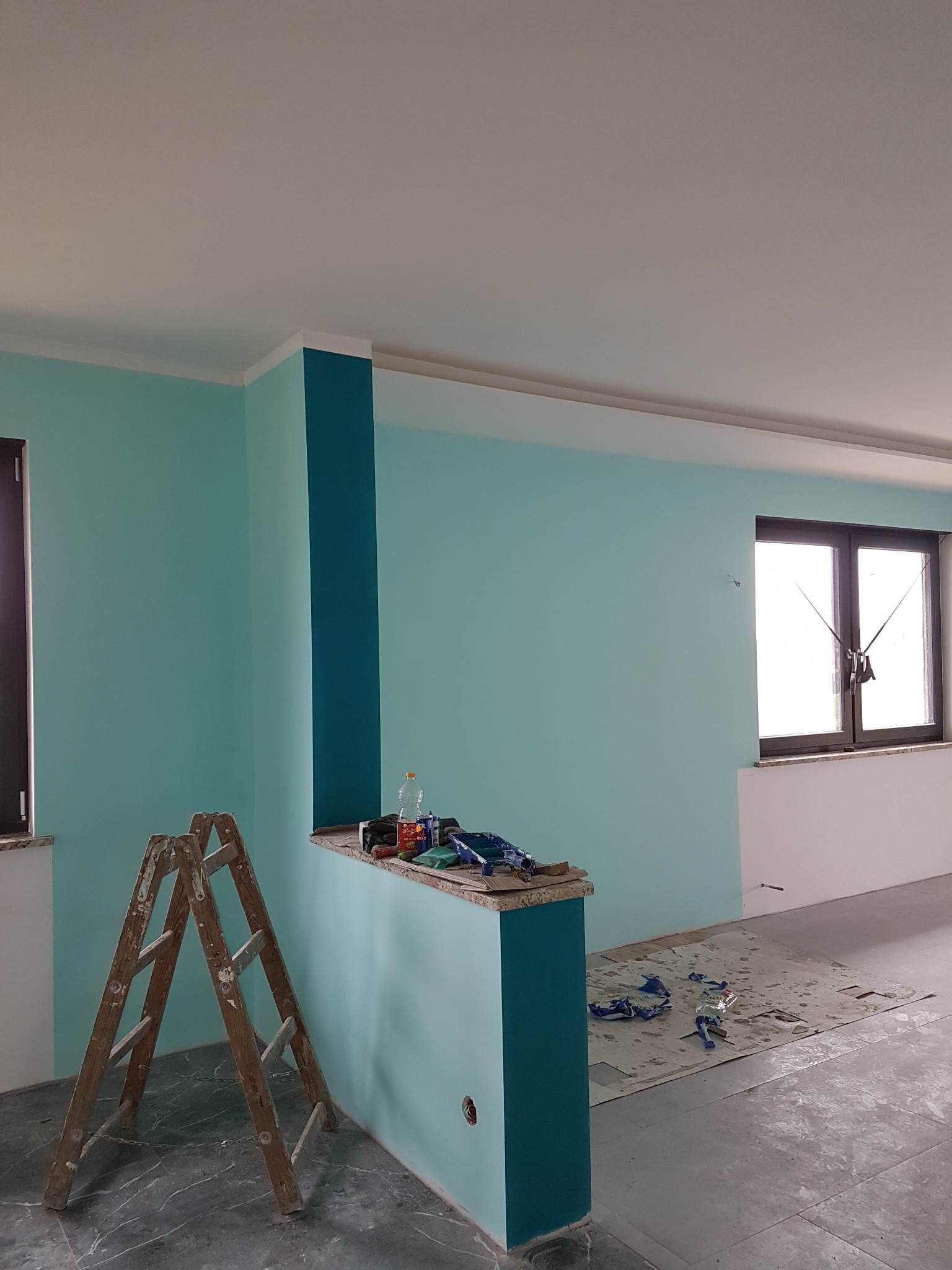 Świeżo pomalowane wnętrze z turkusowymi ścianami, ciemniejszym pionowym pasem, drewnianą drabiną, zabezpieczoną podłogą i oknem z ciemną ramą.
