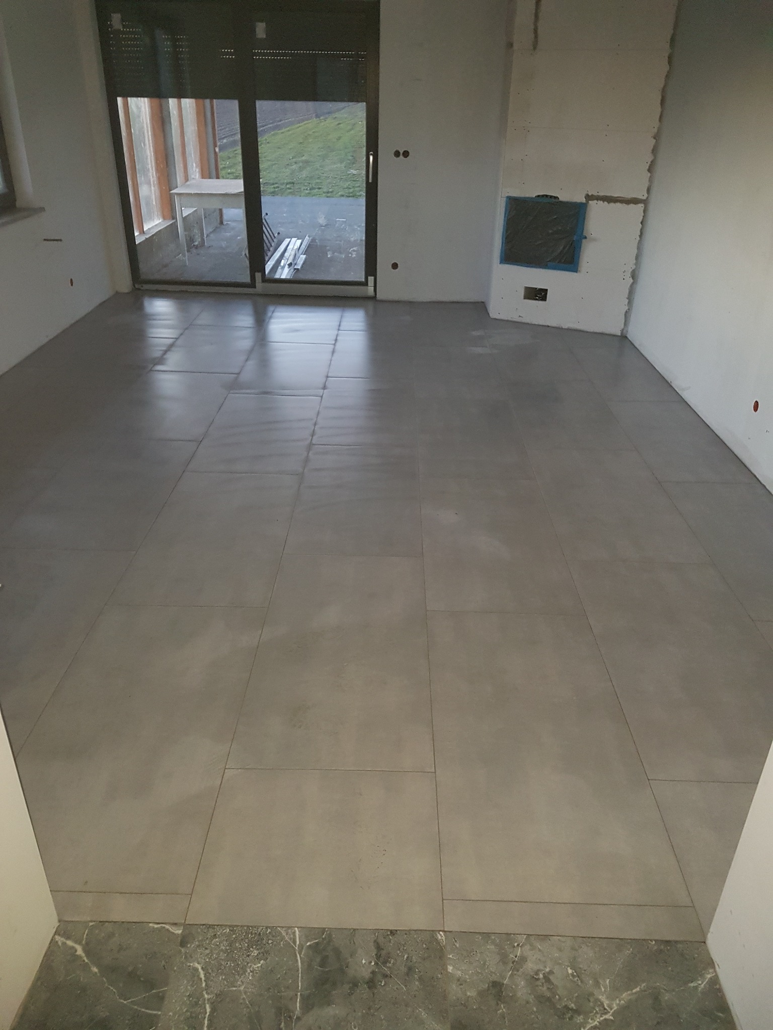 Ułożone szare płytki podłogowe w surowym wnętrzu z widokiem na zewnątrz przez duże przesuwne drzwi balkonowe.