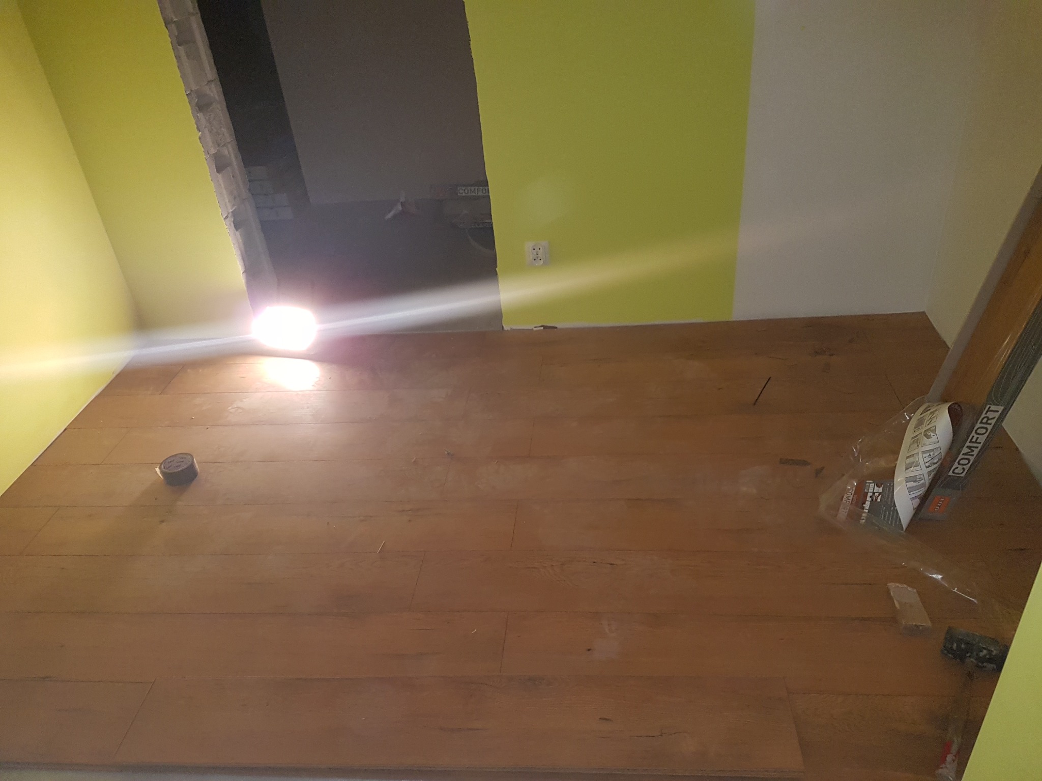 Fragment pomieszczenia w trakcie prac wykończeniowych: ułożone panele podłogowe w odcieniach brązu, widoczne narzędzia i opakowania, ściany w kolorze żółtym i białym, przejście do innego...