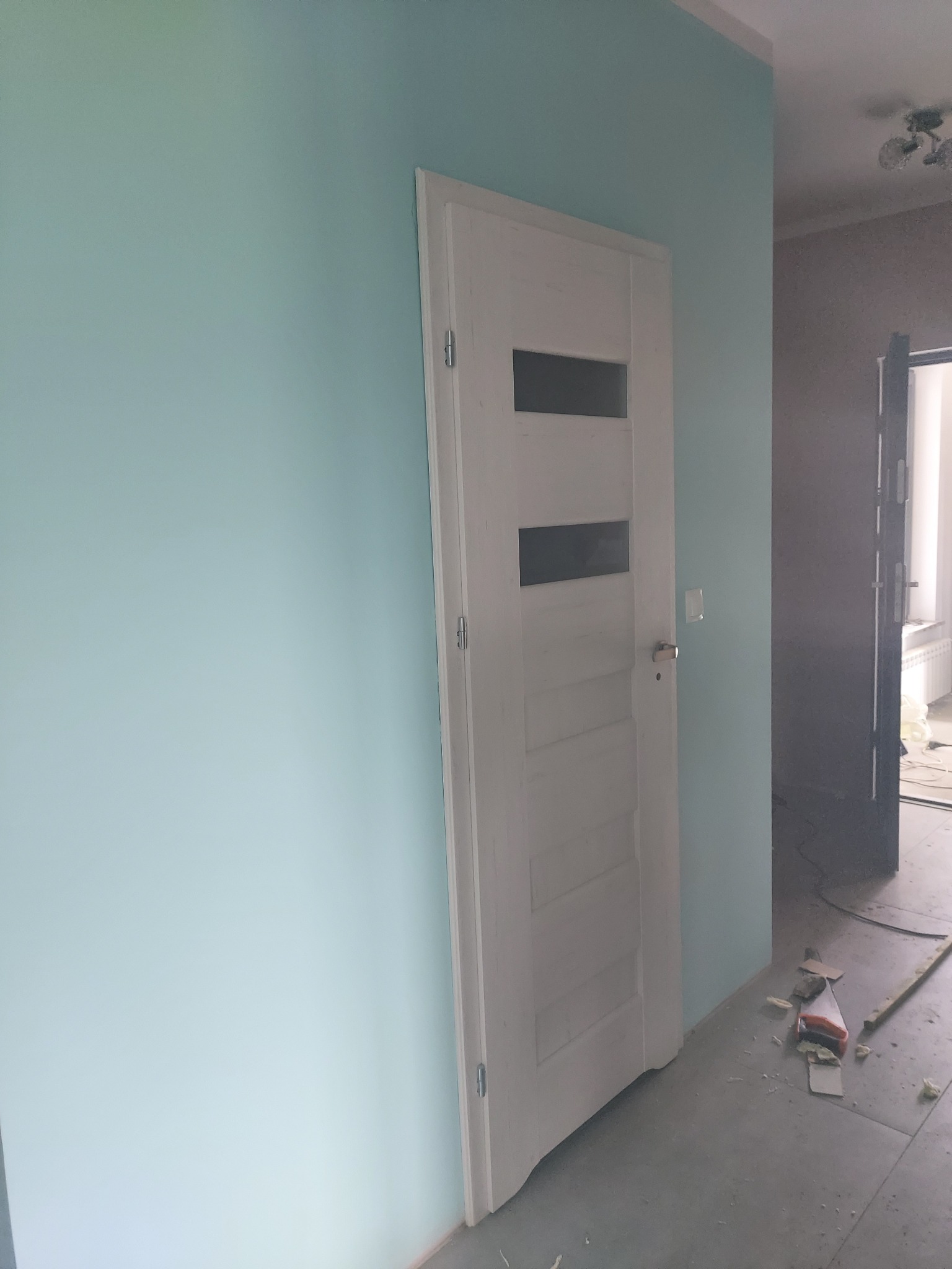 Świeżo pomalowana na błękitno ściana z zamontowanymi białymi drzwiami z trzema poziomymi przeszkleniami, widoczne ślady po remoncie na podłodze.