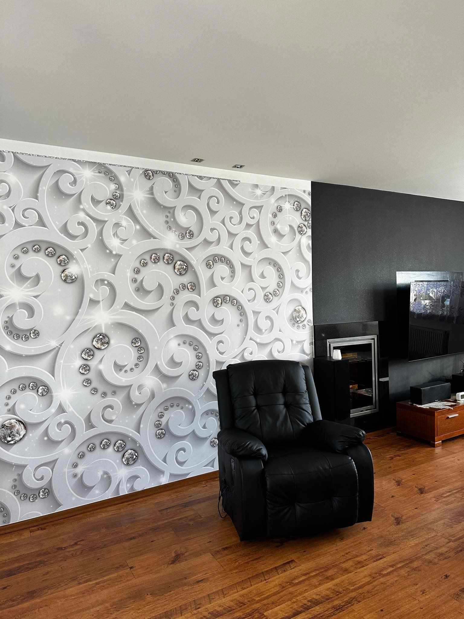 Ściana z dekoracyjną tapetą w srebrne, ornamentowe wzory z kryształkami, sąsiadująca z czarną ścianą w salonie z drewnianą podłogą i czarnym skórzanym fotelem.