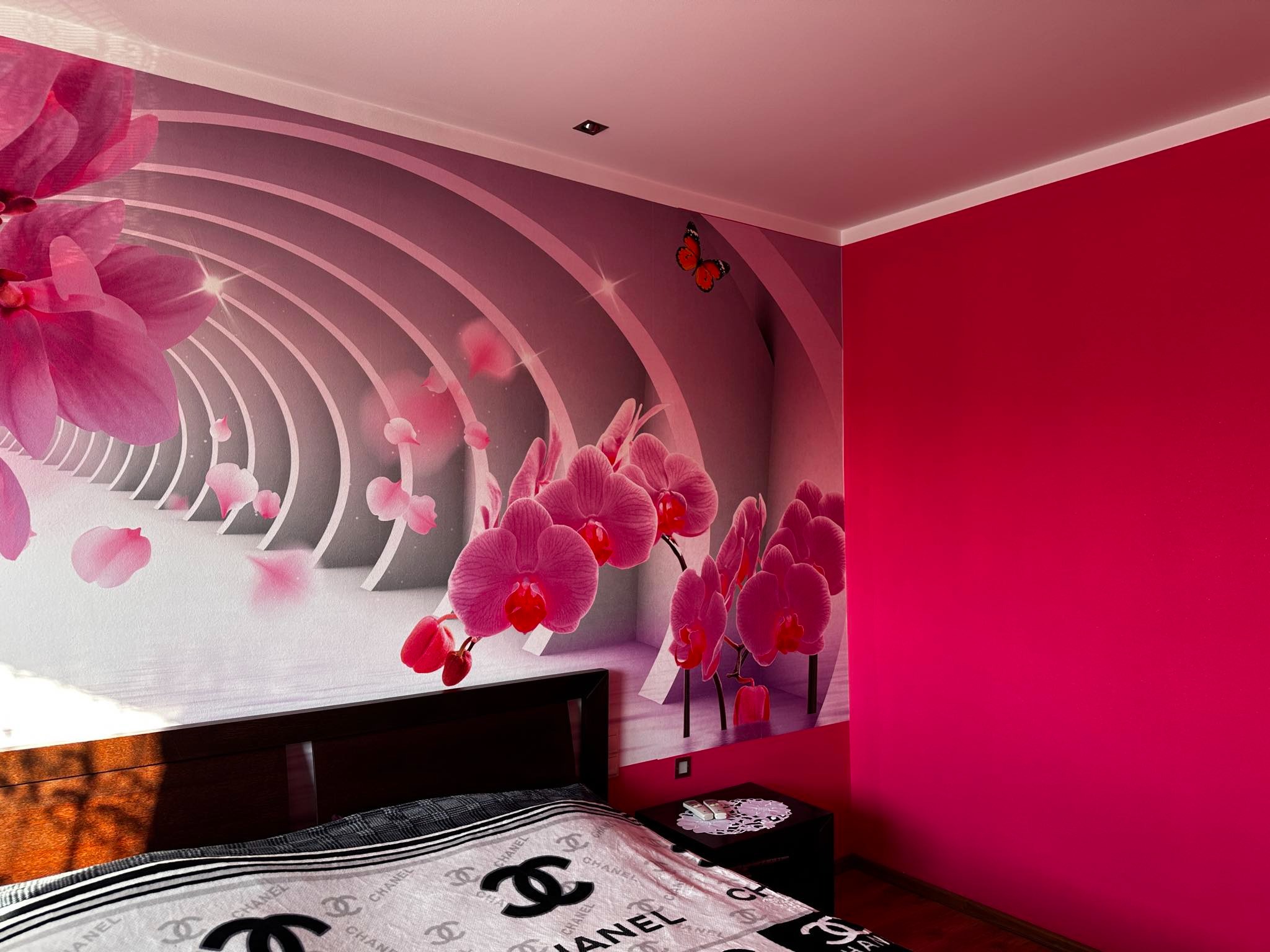 Pokój z fototapetą z motywem orchidei i tunelu, ściana pomalowana na intensywny róż, łóżko z narzutą w logo Chanel.