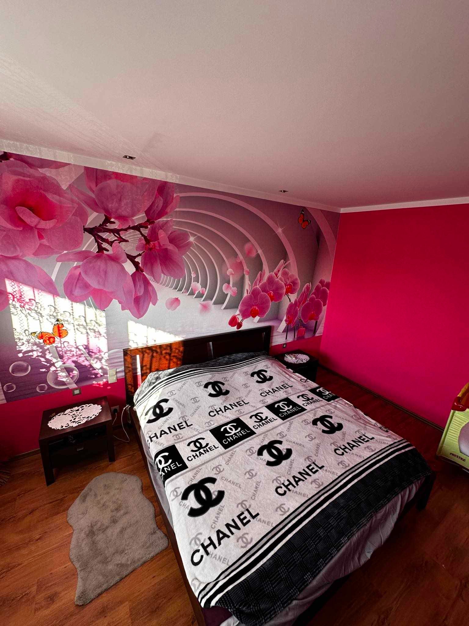 Pokój z fototapetą z motywem kwiatowym i łóżkiem z pościelą marki Chanel, na podłodze dywanik imitujący skórę owcy, obok łóżka stolik nocny.