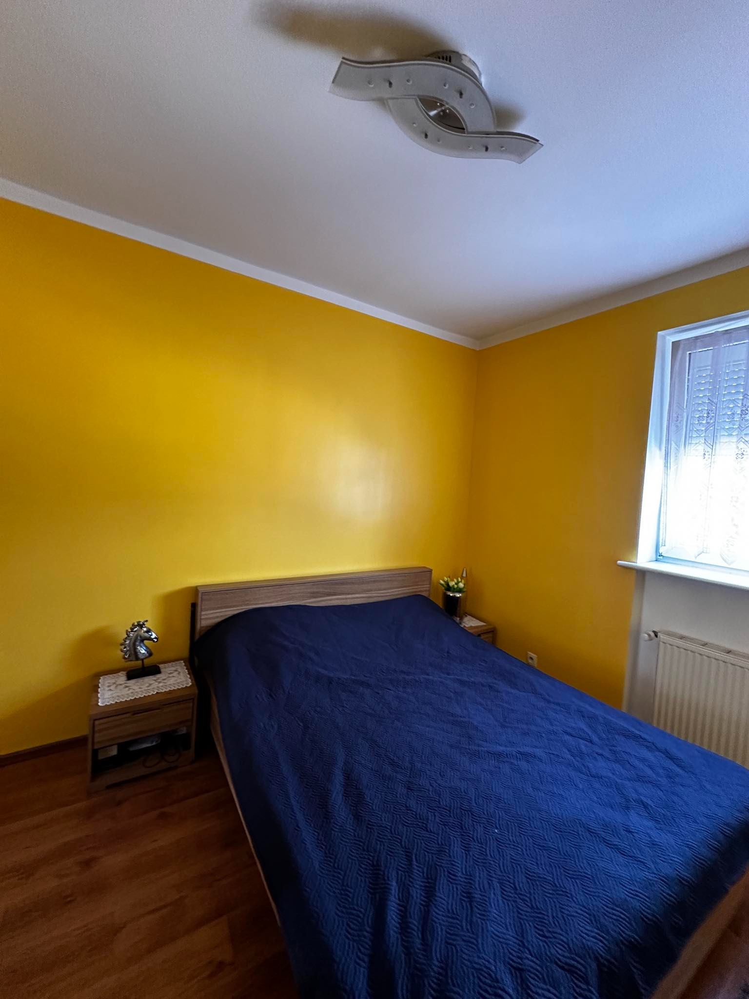 Sypialnia z intensywnie żółtymi ścianami, drewnianą podłogą i niebieską narzutą na łóżku; na szafce nocnej dekoracja w kształcie konia.