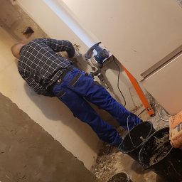 REMS & STEL DYNAMICS - Pracownik w niebieskich spodniach i kraciastej koszuli używa młota udarowego do kucia ściany podczas remontu, widoczne wiadra, poziomica i worek z zaprawą.